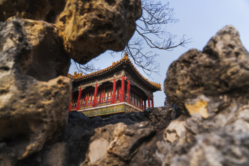 View of a traditional Chinese building, framed by rough, textured rocks, its red pillars and golden roof standing out against the pale sky, Beijing, Beijing, China.