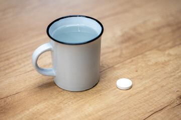 Effervescent vitamin tablet next to a mug of water on a wooden table. Fast-dissolving supplement for quick absorption, hydration support, digestion aid, and daily vitamin intake.