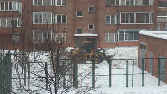 Yellow front loader clearing snow near a brick residential building and fenced playground in a snowy winter landscape
