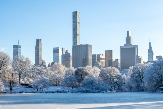 New York City winter scene in Central Park with frozen lake and snow-covered trees, Midtown Manhattan skyscrapers rising in the background. Peaceful, cold urban landscape in NYC, USA