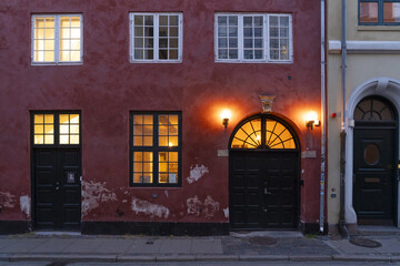 Copenhagen, Denmark - 30 September 2025: View of a dark red building facade illuminated by warm light spilling from windows and doorways, contrasting with the cool grey pavement below.