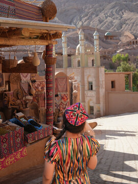 Turpan, China - 25 June 2025: View of a woman in traditional attire standing amidst colorful textiles with the Emin Minaret rising in the background.