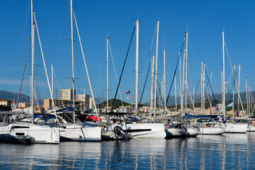 Obraz premium Wide view of sailboats docked side by side in a marina with tall masts, city buildings, and mountains in the background.