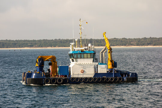 Multipurpose offshore support vessel with yellow cranes sailing at sea, providing maritime logistics and construction services along a coastal landscape.