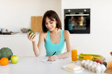 A young woman smiles as she writes notes in a bright kitchen filled with fresh ingredients like avocado, eggs, and vegetables. A glass of juice sits beside her on the counter.