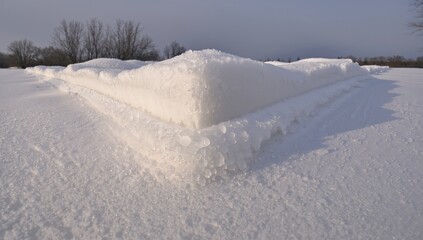 Showing triangular layered snow and ice ridge casting soft shadows in rural field, with tracks