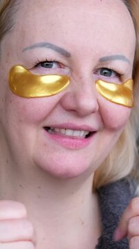 Adorable curly woman glancing straight on camera. Happy woman smiling and using gold eye patches with collagen, peptide, ceramide and hyaluronic acid, derma oil. Dermatology, healthy skin concept.