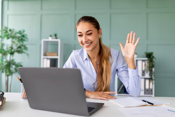 Cheerful woman sits at her desk, engaging in a virtual meeting on her laptop. She smiles and waves to the webcam, teaching students online from her home.
