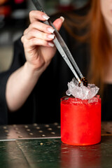 Bartender garnishing vibrant red cocktail on crushed ice with tweezers at a bar counter. Close-up shot, moody lighting