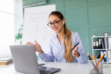 A female teacher conducts an engaging online class. She is using a laptop for a video call with students while wearing wireless earbuds. She explains concepts clearly and gestures to help.