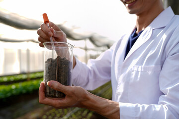 Asian man in lab coat and safety glasses putting soil sample into test tube doing research in greenhouse