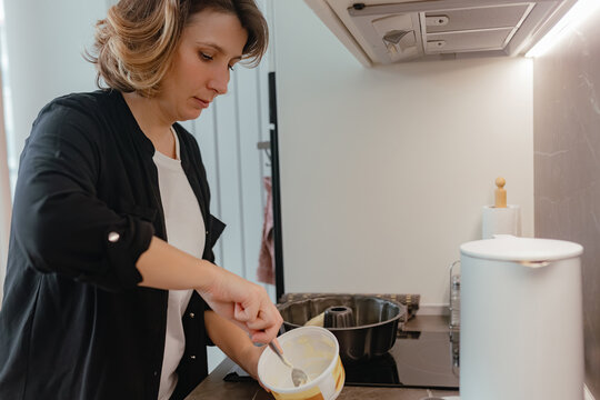 Adult woman scoops margarine from yellow container while preparing mixture near stovetop area inside modern bright kitchen under warm illumination.