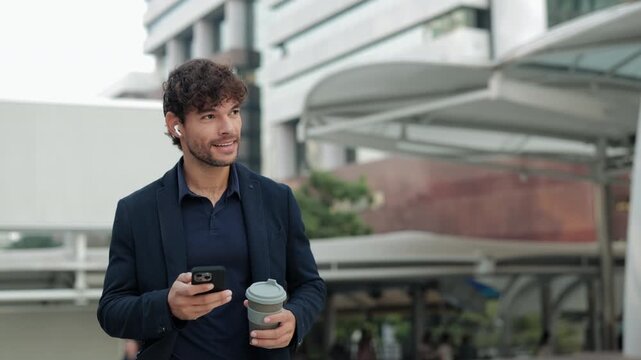 Portrait of young Hispanic office employee drinks coffee looking at smartphone on urban street. Latin man entrepreneur scrolls internet walking to workplace in downtown district
