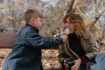 Child holds ice cream near woman during autumn stroll, casual clothing, warm sunlight across bench, yellow foliage around trees, relaxed interaction, natural outdoor lifestyle scene.