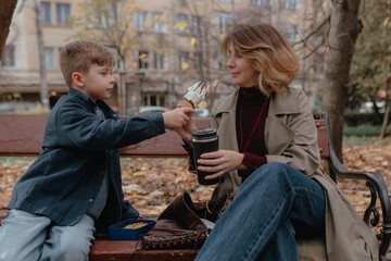 Boy extends ice cream toward woman during autumn walk, yellow foliage around park, natural textures, casual outfits, relaxed facial focus, stainless cup near bench, soft daylight across scene.