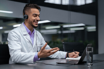 A doctor in a white coat uses a laptop and headset to conduct a virtual consultation. He interacts with a patient, taking notes during the session in a modern office.