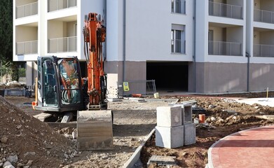 Front view of  compact excavator  with a  modern residential building under construction on behind. 