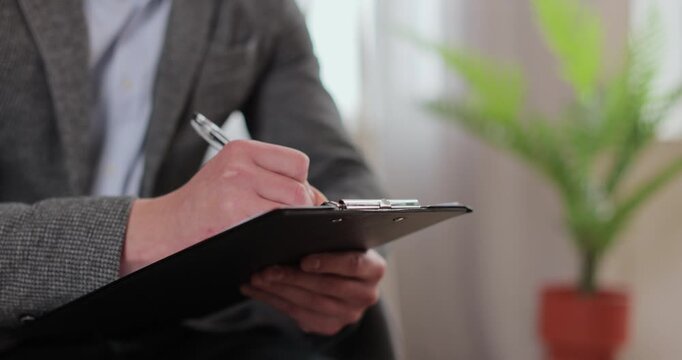 Close up man psychologist writing observation notes during therapy session with patient sitting beside in clinic room for mental health counseling support.