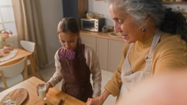 Medium close up POV of elderly Caucasian woman vlogging on camera with granddaughter in apron while tasting freshly baked loaf cake at home kitchen