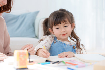 Cheerful Asian mother and little daughter enjoying creative activity together at a bright, cozy white table in the living room. Focus on happy family bonding, learning, and childhood development.