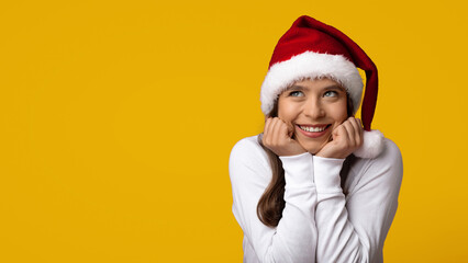 A happy girl wearing a Santa hat smiles excitedly, resting her chin on her hands. The bright yellow background adds a festive touch, capturing the spirit of the holiday season.