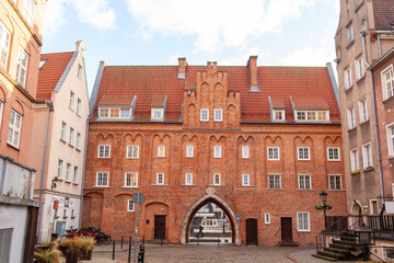 Fototapeta premium A brick building stands in a quiet urban square under a clear sky. The street is empty with a few bicycles parked nearby. Chlebnicka Gate in Gdansk, Poland