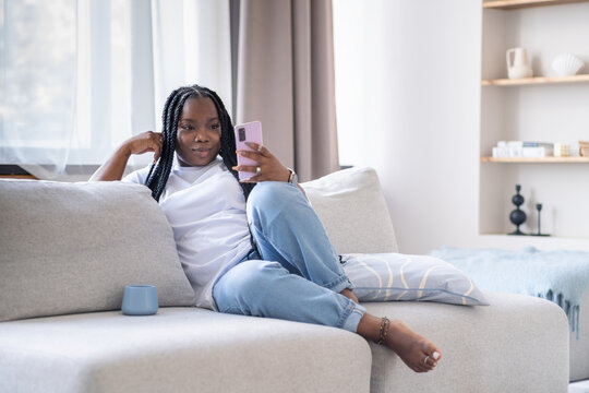Excited happy young black woman holding smart phone device sitting on sofa at home - Happy satisfied female looking at mobile smartphone screen- Technology concept