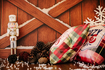 Classic Christmas Still Life Featuring a White and Gold Nutcracker Figure Standing Beside a Pile of Pinecones and Plaid Pillows, One Reading 'Cozy Christmas', Set Against a Rustic Wooden Background