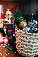 Close-up of a Modern Purple and Gold Nutcracker King Figurine Standing Beside a Woven Basket Filled with Shiny Silver and Gold Christmas Ornaments and Faux Pine Needles on a Wooden Surface