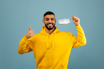 Naklejka premium A man wearing a yellow hoodie smiles broadly while waving a mask in one hand and giving a thumbs up with the other. The background is light blue, enhancing his cheerful demeanor.