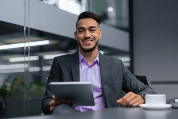 Fototapeta premium A man is sitting at a desk in a modern office environment. He holds a tablet and smiles while wearing a suit. A cup is placed on the table next to him.