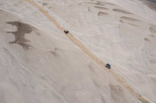 Aerial view of two vehicles kicking up dust as they navigate a dirt path through a vast expanse of sandy terrain, Sariakandi, Rajshahi Division, Bangladesh.
