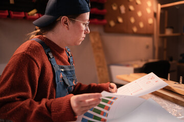 Female carpenter studying documents, reports and charts while working late evening in workshop, small business and craftsmanship concept