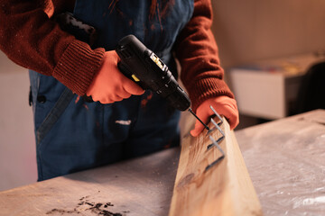Carpenter drills wooden plank with electrical drill. Woman Joiner working in her carpentry workshop.