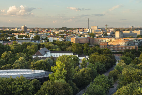 View of lush green treetops concealing buildings below a skyline with a tall spire, bathed in the warm glow of the setting sun, Tiergarten, Berlin, Germany.