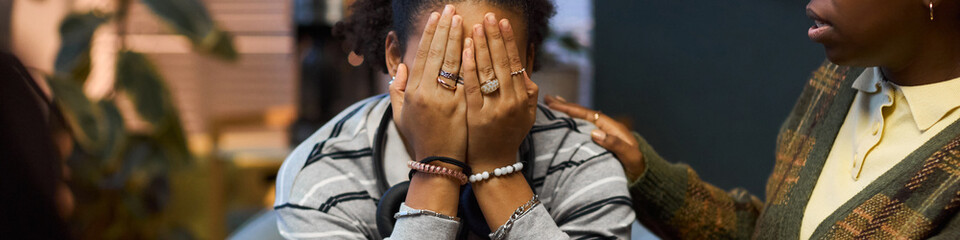 Fototapeta premium Black woman comforting young Black woman sitting with head in hands, offering support in school setting, both wearing casual clothing, emotional moment between adults