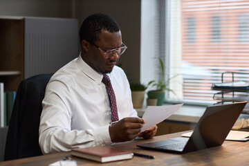 Middle aged Black man sitting at desk reading document, wearing glasses and formal attire, working in office with laptop and notebook, school principal reviewing paperwork © DragonImages
