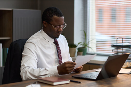 Middle aged Black man sitting at desk reading document, wearing glasses and formal attire, working in office with laptop and notebook, school principal reviewing paperwork