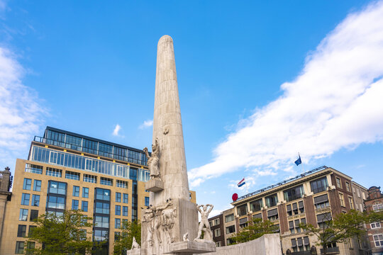 View of a towering white monument, etched with figures, rises towards the vibrant blue sky, framed by classic and modern buildings, Amsterdam, North Holland, Netherlands.
