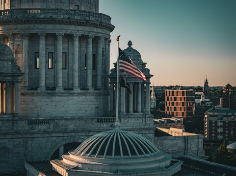 Aerial view of the Rhode Island State House, its dome glowing softly against the skyline, with the American flag waving proudly above, Providence, Rhode Island, United States.