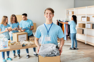 In a charity center, a male volunteer in uniform holds a cardboard box filled with clothes for donation.