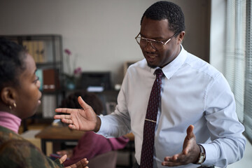 Middle aged Black man gesturing while speaking to young Black woman in office setting,...