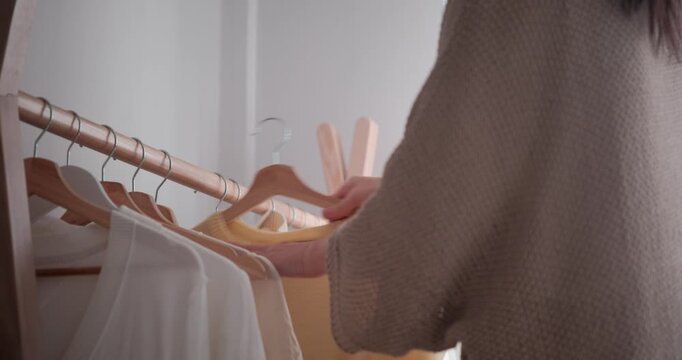 Rear view of woman choosing clothes from rack in bedroom. Female hands sorting through hangers with sweaters and picking yellow one. Getting ready routine at home.