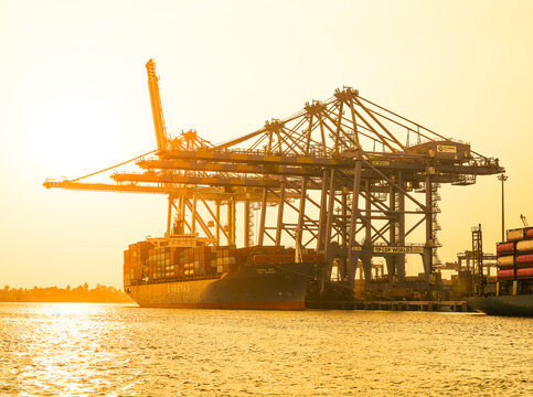 View of a massive cargo ship dwarfed by towering cranes under a golden sky, docked at the bustling port in Kochi, Kerala, India.