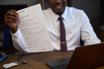 Middle aged Black man smiling while holding up graded math test with visible mark, sitting at desk with laptop, representing school principal reviewing student achievement