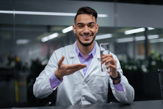 A man in a lab coat smiles while holding a clear glass of water. He gestures with his other hand as he shares information in a modern office space with plants and glass walls.