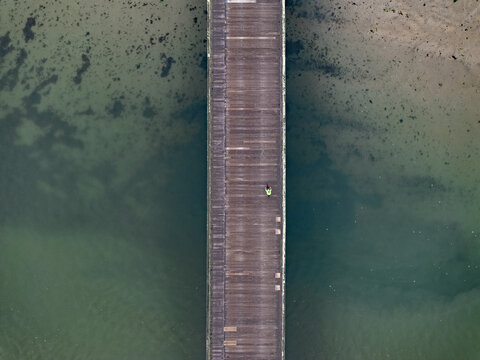Aerial view of a weathered wooden pier stretching over the shimmering, shallow waters, a lone figure punctuating the tranquil scene, Duxbury, Massachusetts, United States.