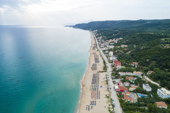 Aerial view of the turquoise sea kissing the golden sands of the beach, meeting the lush green forests and scattered houses, Paralia, Preveza, Greece.