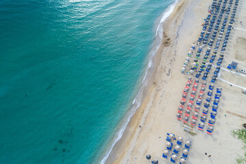 Aerial view of turquoise waves crash onto the golden sands where colorful umbrellas and sunbeds line the shore, Paralia, Preveza, Greece.