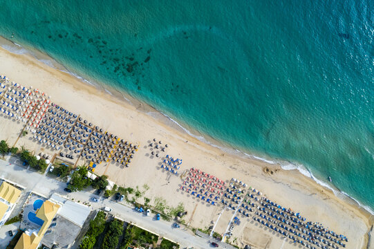 Aerial view of sun-kissed beach where rows of colorful umbrellas punctuate the golden sand meeting the turquoise Ionian Sea, Paralia, Preveza, Greece.